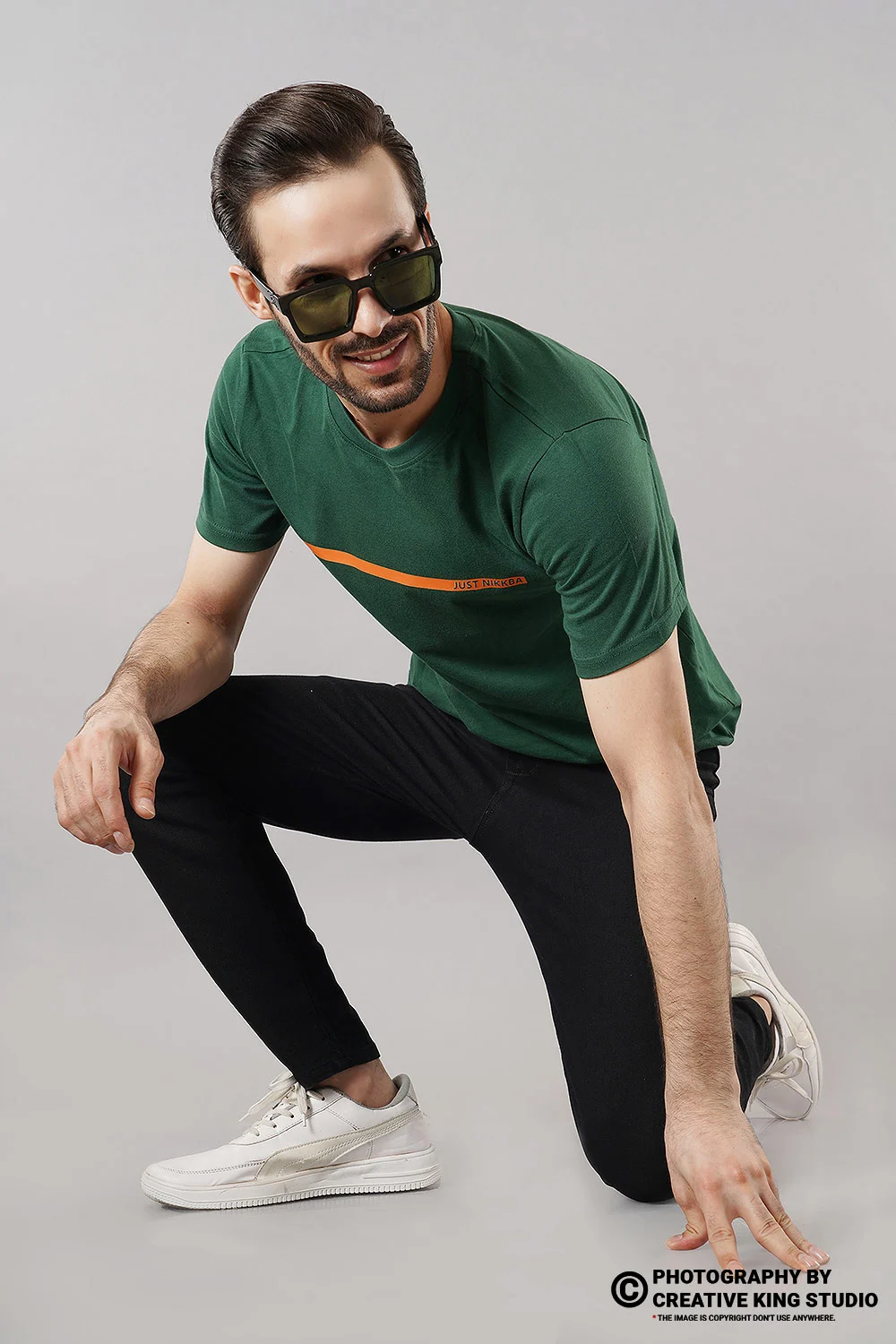 Male model in a green crew-neck t-shirt and black joggers on a grey studio backdrop, an on-model apparel catalogue pose showing fit and length