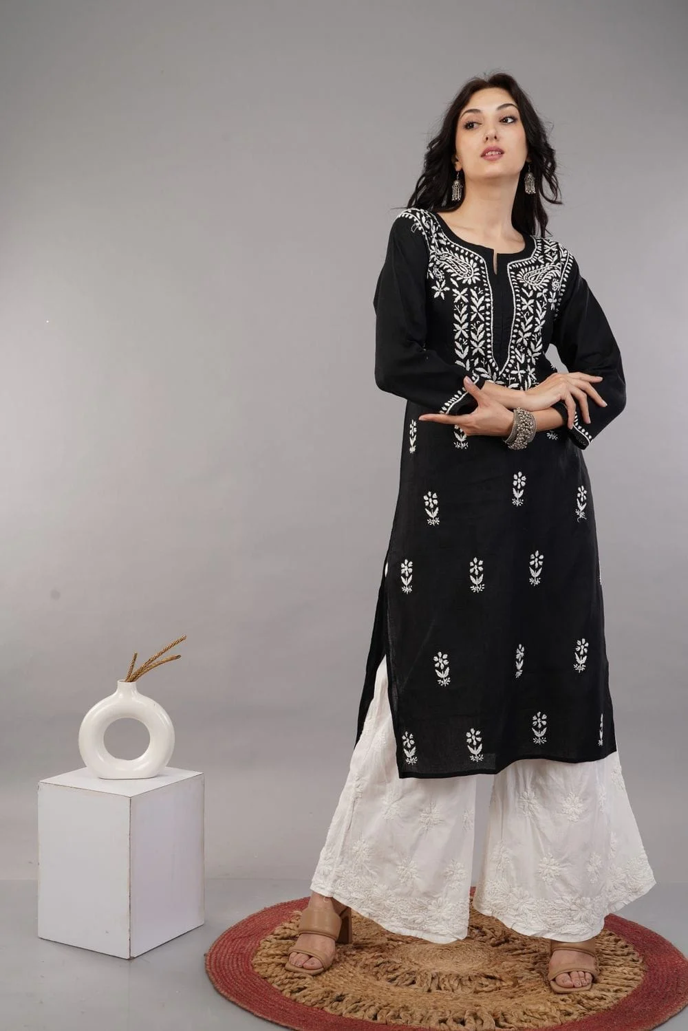 Black embroidered kurta set styled with a vase, plinth and jute rug on a grey set, suited to a secondary lifestyle frame rather than the main image