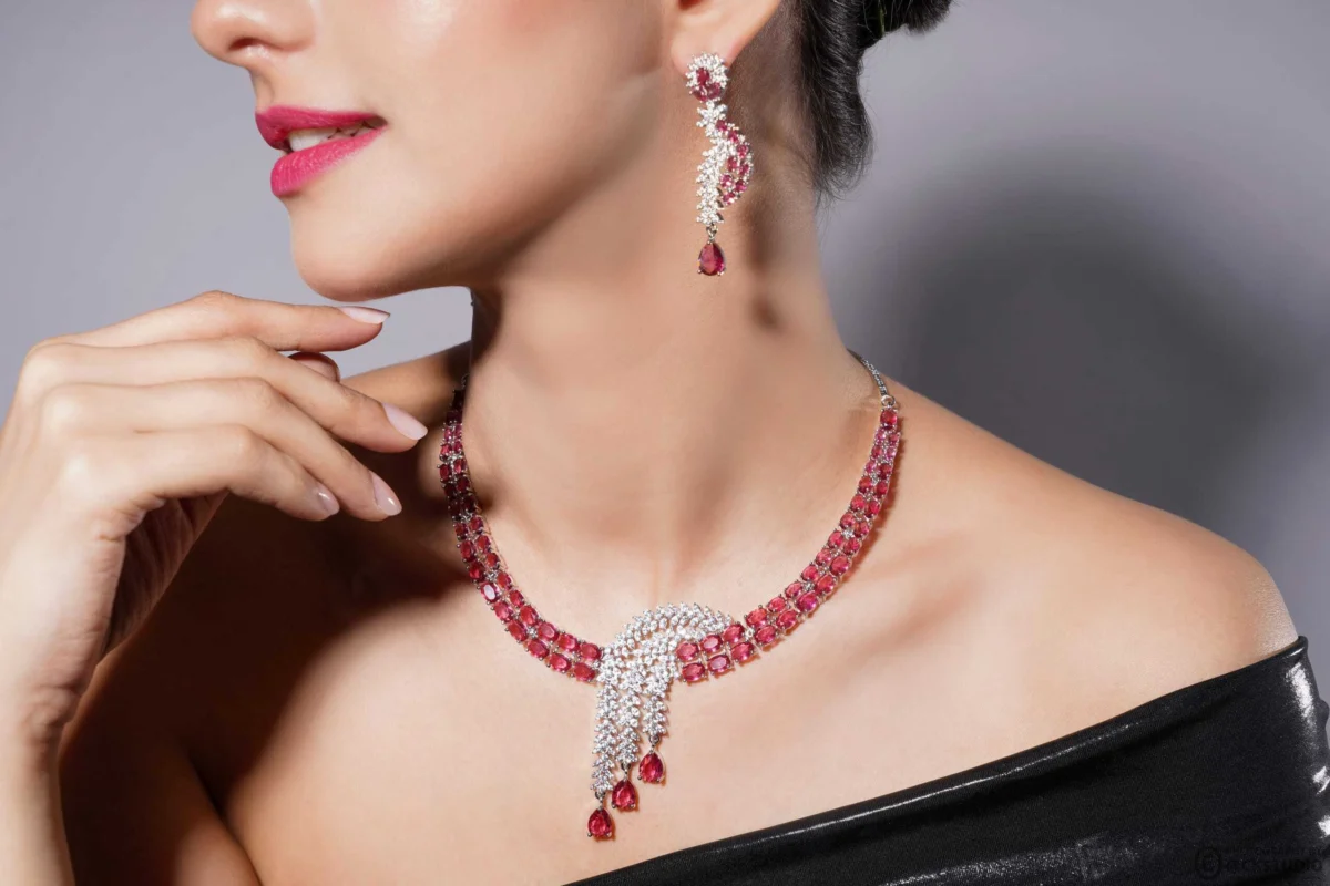 Model in a black off-shoulder top wearing a ruby and diamond necklace with matching drop earrings against a soft grey backdrop, a lifestyle image for a jewellery set