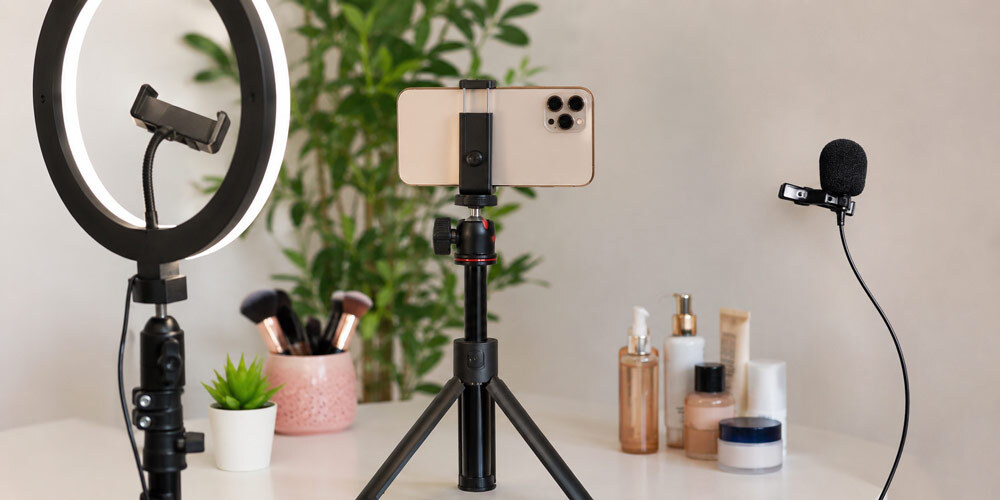 Compact UGC creator equipment setup featuring smartphone on adjustable tripod, ring light with phone holder, clip-on lavalier microphone and beauty product props arranged on minimalist white desk, professional home studio workspace for short-form video production