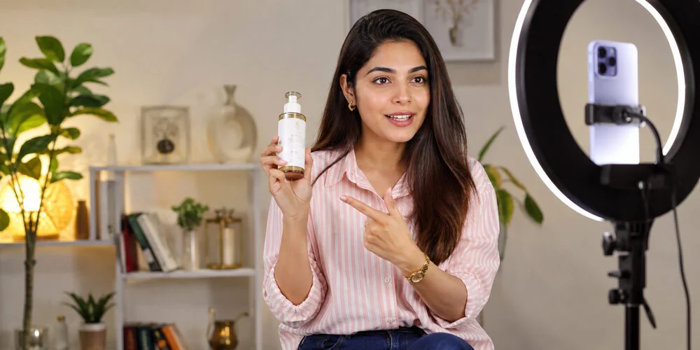 Indian female creator in a striped shirt holding a skincare bottle and pointing at it while recording a vertical product review on a smartphone mounted in a ring light, home-style shelf backdrop
