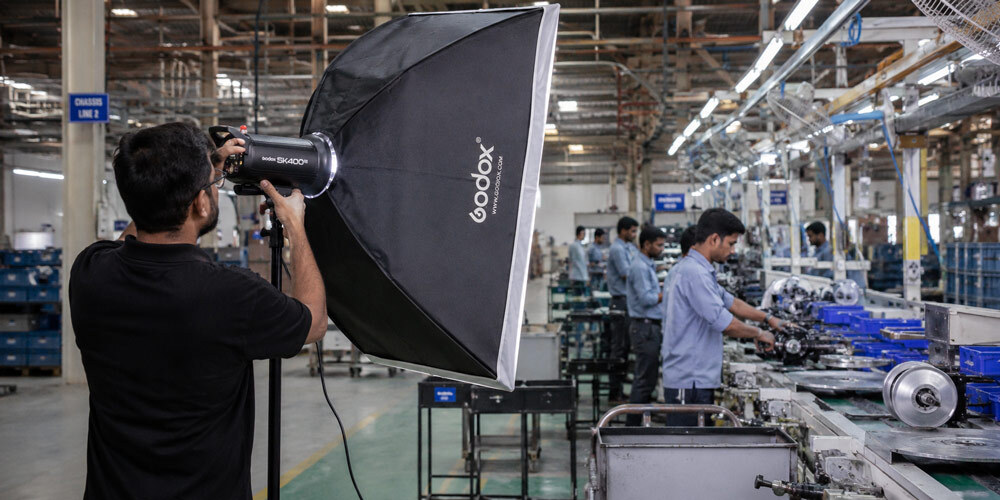 Professional industrial photographer setting up large softbox lighting to capture factory production line operations with clear focus on quality manufacturing detail in an Indian plant