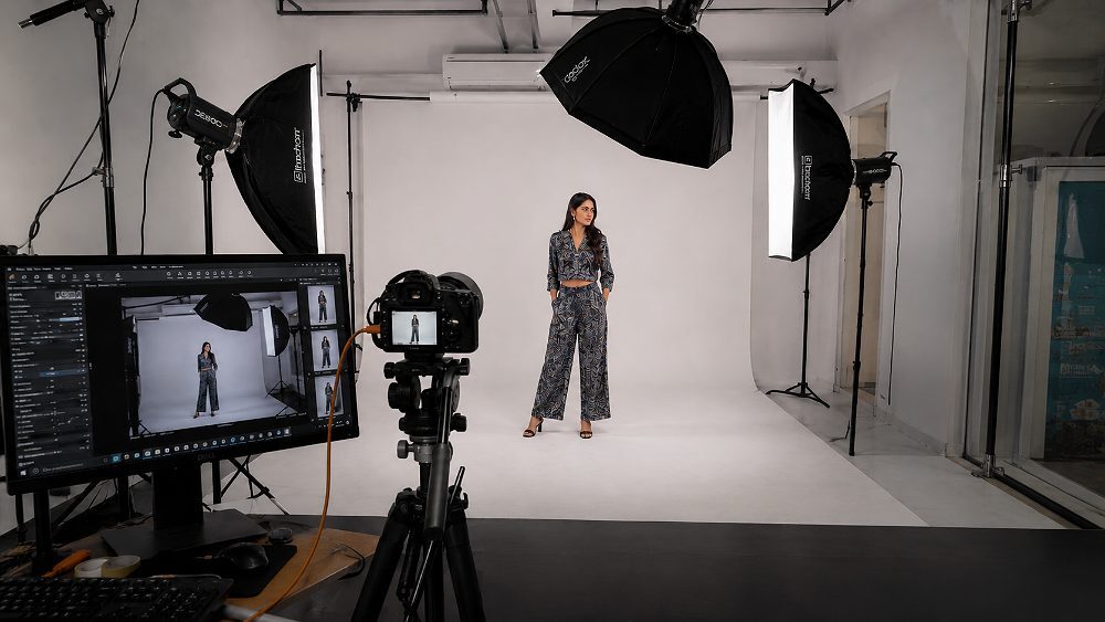 Behind the scenes tethered capture workflow at Indian standard ecommerce photography studio showing a full frame mirrorless camera on tripod aimed at a woman model in a co-ord set on cyclorama with two strip softboxes and one octabox rigged on Godox and Elinchrom strobes plus a colour calibrated monitor running Capture One and barcoded SKU rack in the foreground for multi-marketplace clothing catalogue shoot