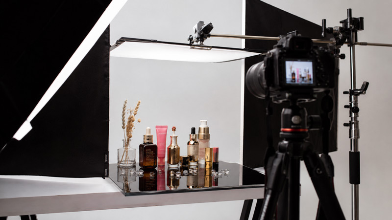 Behind the scenes photograph of Ckstudio beauty product photography tabletop with cosmetic serum bottles and lipsticks arranged under reflection-controlled softbox lighting with black flags on beauty photography set