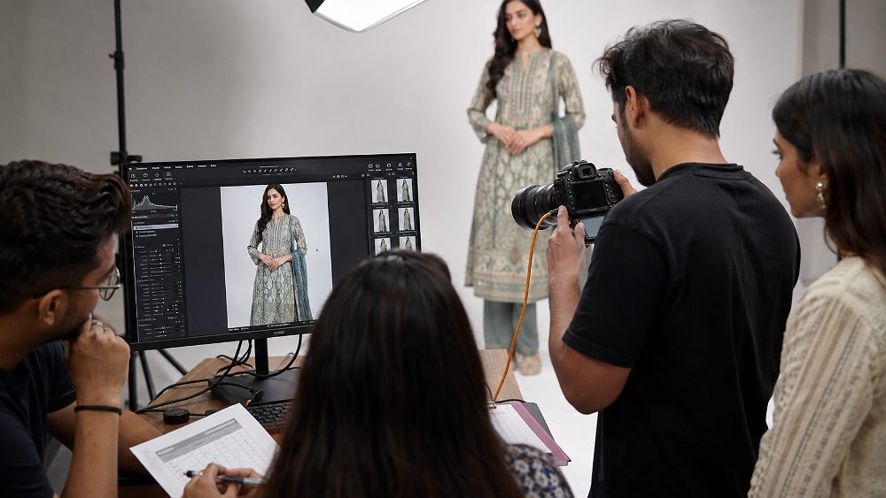 Tethered shooting workflow during Fashion Catalogue Shoot at Indian standard ecommerce photography studio showing photographer holding DSLR camera connected via USB cable to twenty-seven-inch calibrated monitor displaying live preview of model in kurta, brand representative reviewing frame and stylist noting continuity in shared document beside the tethered station