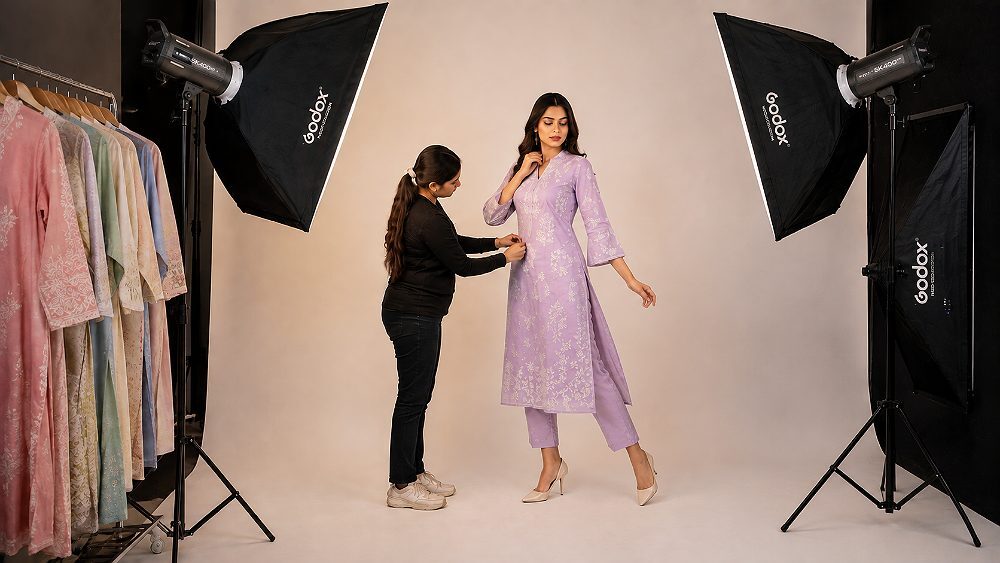 Behind the scenes of a Fashion Catalogue Shoot at an Indian standard ecommerce photography studio with model posing under three-point lighting on seamless ivory backdrop, stylist adjusting garment fit and photographer reviewing tethered frames on monitor for D2C fashion brand production