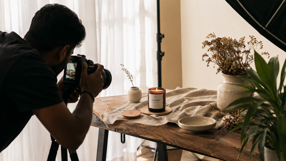 Behind the scenes photograph of Ckstudio lifestyle product photography set with scented candle on wooden coffee table styled with linen fabric and ceramic props under soft window daylight for D2C brand hero imagery