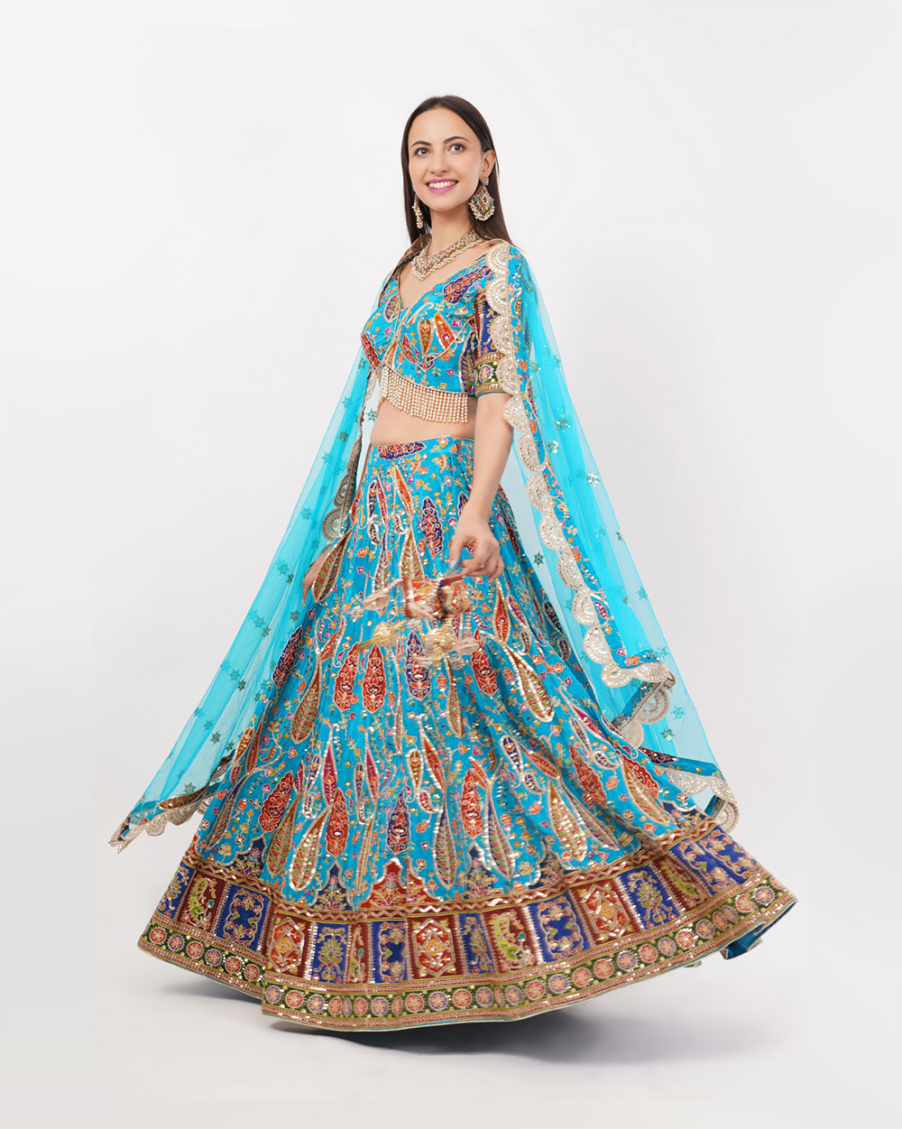 Indian female fashion model wearing a vibrant turquoise blue embroidered lehenga choli with multicolour traditional motifs, matching sheer dupatta, statement jewellery and clutch, posing against a clean white studio background beside a decorative floor vase during professional ethnic wear fashion photography